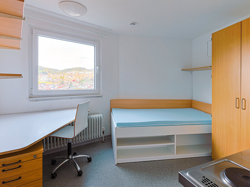 Helles Zimmer mit Bett, Schreibtisch, Stuhl und Fenster mit Blick nach draußen.