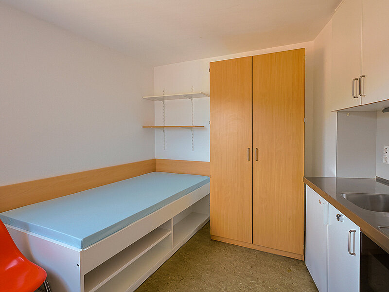 Zimmer mit Bett, Schrank, Regal und integrierter Küchenzeile.